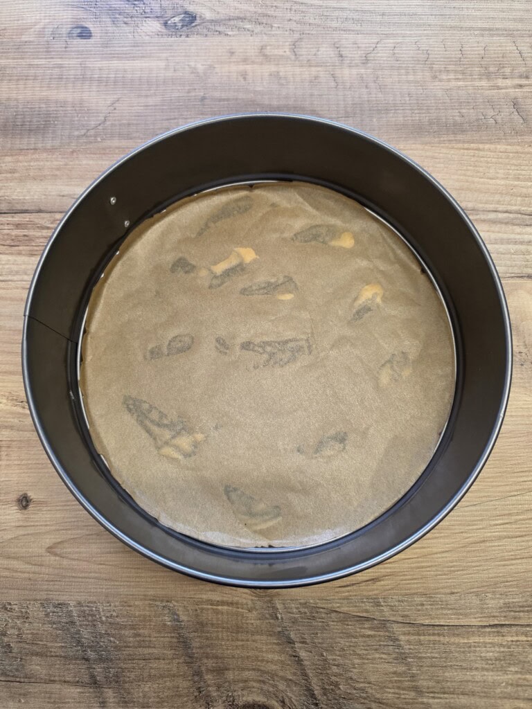 A round springform cake tin lined with baking paper on the bottom, placed on a wooden surface. The baking paper is slightly wrinkled and has a few areas with visible grease marks.