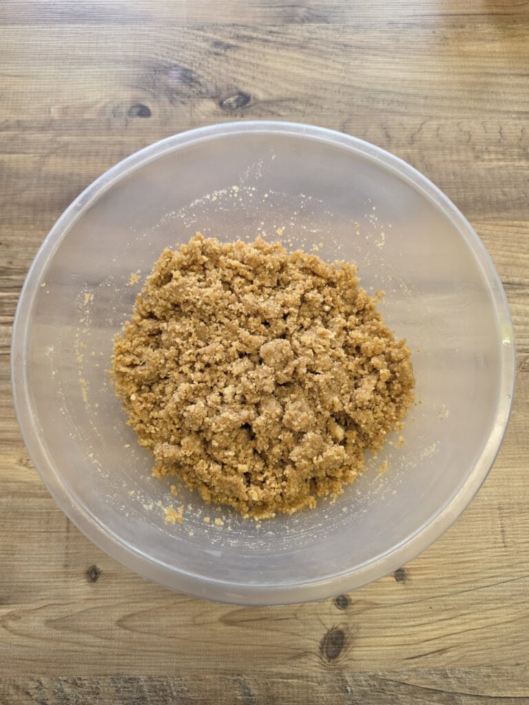 A clear mixing bowl with crumbly, light brown dough or mixture inside, placed on a wooden surface.