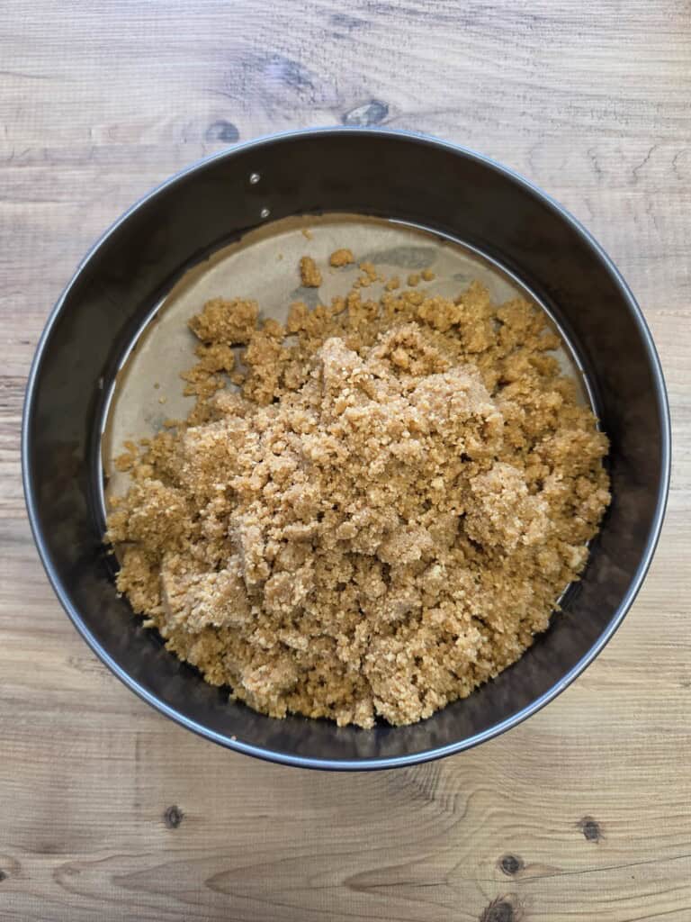 A round baking tin containing a pile of crumbly, light brown biscuit or digestive biscuit base mixture on a wooden surface.