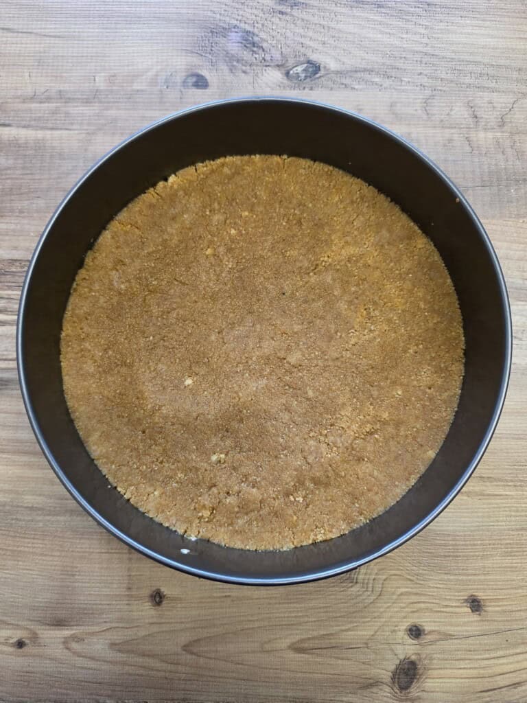 A round baking tin filled with an even layer of pressed digestive biscuit base sits on a wooden surface.