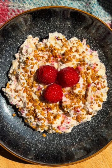 A black plate holds creamy cranachan mixture topped with toasted oats, honey and three raspberries, set on a wooden surface with a colourful fabric in the background.