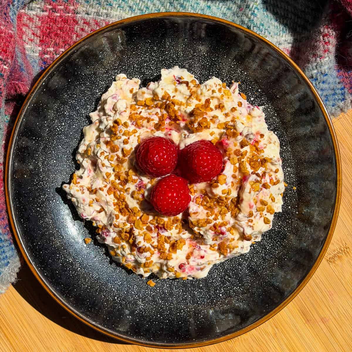 A black plate holds creamy cranachan mixture topped with toasted oats, honey and three raspberries, set on a wooden surface with a colourful fabric in the background.