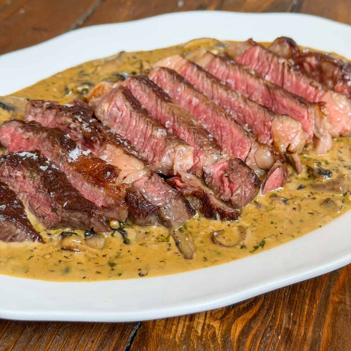 Sliced medium-rare steak sits atop a creamy mushroom sauce on a white plate, placed on a wooden table. The inside of the steak is pink and juicy, whilst the outside is seared and browned.