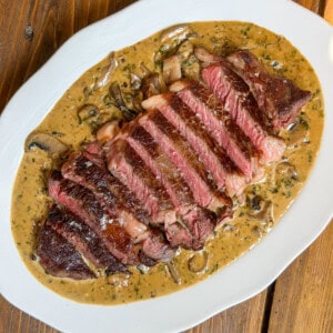 Sliced medium-rare steak served on a white plate, topped with a Diane sauce and garnished with herbs, on a wooden table.