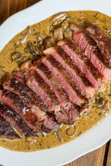 Sliced medium-rare steak served on a white plate, topped with a Diane sauce and garnished with herbs, on a wooden table.