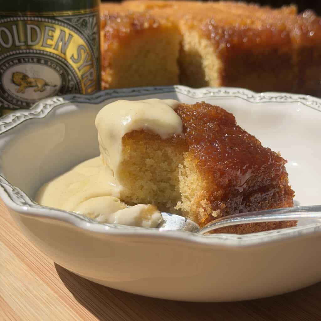 A slice of sticky syrup sponge pudding with golden syrup and creamy custard in a bowl, with a spoon and the rest of the cake and syrup tin in the background.