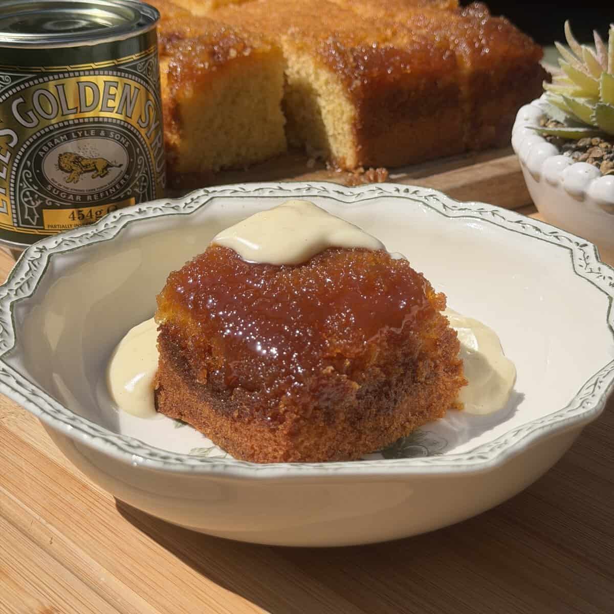 A slice of golden syrup sponge cake with cream sits in a patterned bowl. Behind it are a tin of Lyle’s Golden Syrup, the remaining cake on a board, and a potted succulent. Sunlight brightens the wooden surface.