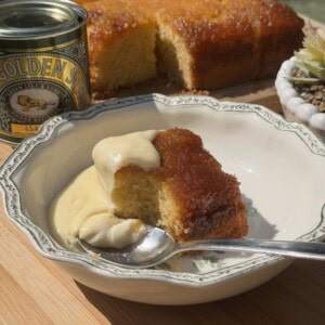 A slice of syrup sponge pudding topped with custard sits in a decorative bowl, with a spoon and a tin of Golden Syrup nearby. The rest of the pudding is in the background.
