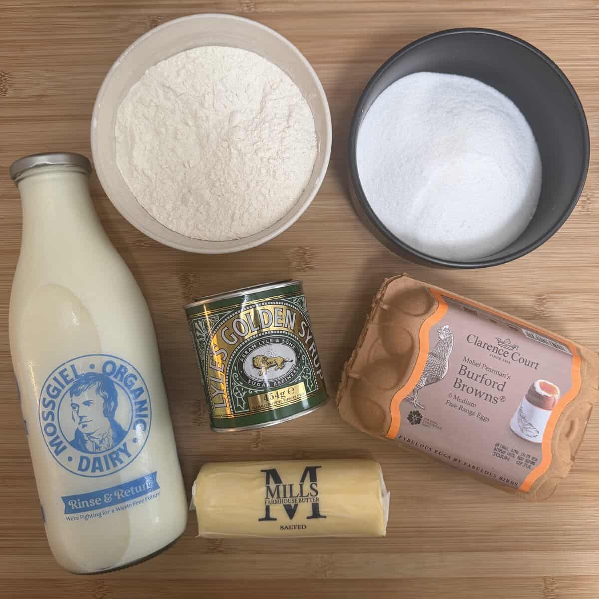 Baking ingredients on a wooden surface: a bottle of milk, a bowl of flour, a bowl of sugar, a tin of golden syrup, a pack of salted butter, and a carton of Burford Brown eggs.