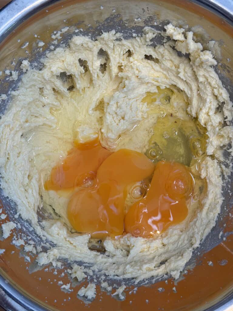 Four raw eggs are cracked into a bowl of creamed butter and sugar, ready to be mixed. The mixture is in a metal bowl, and the eggs are sitting on top of the partially blended ingredients.