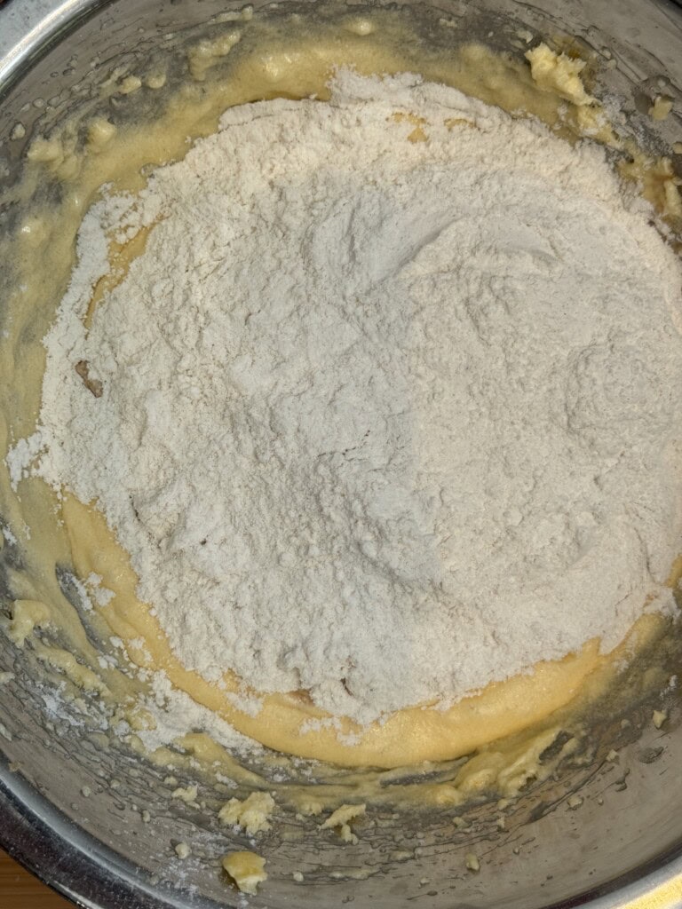 A metal bowl containing a yellow batter with a pile of white flour on top, ready to be mixed together for baking.
