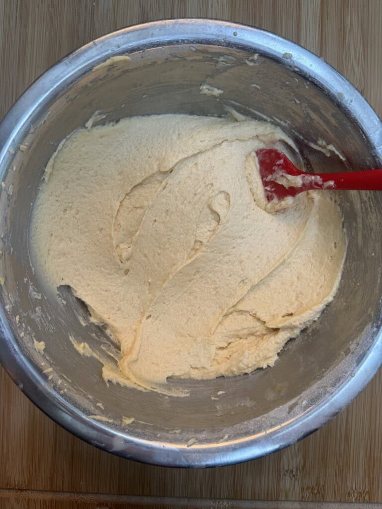 A stainless steel bowl filled with creamy yellow cake mixture on a wooden surface. A red spatula rests inside the bowl, partially covered with mixture.
