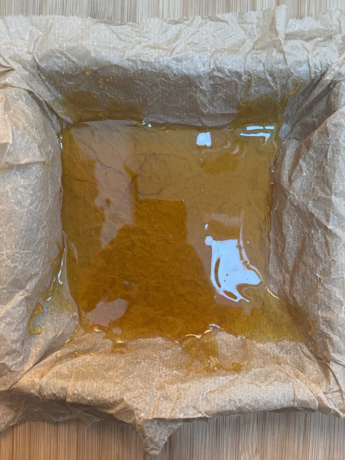 A square baking dish lined with baking parchment holds a layer of golden liquid, possibly melted butter or syrup, on a light wooden surface.