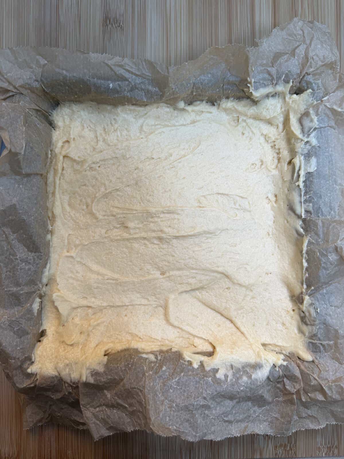 Square baking tin lined with greaseproof paper, filled with an even layer of light-coloured batter, ready to be baked. The tin sits on a wooden surface.