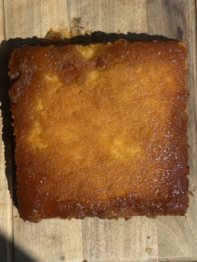 A golden-brown, rectangular cake with a shiny caramelised top sits on a wooden surface, lit by sunlight.
