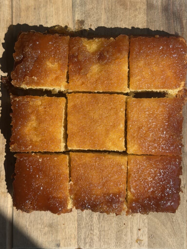 Nine pieces of golden-brown syrup sponge cake with a shiny, caramelised top are arranged neatly on a wooden surface, with sunlight highlighting their texture.