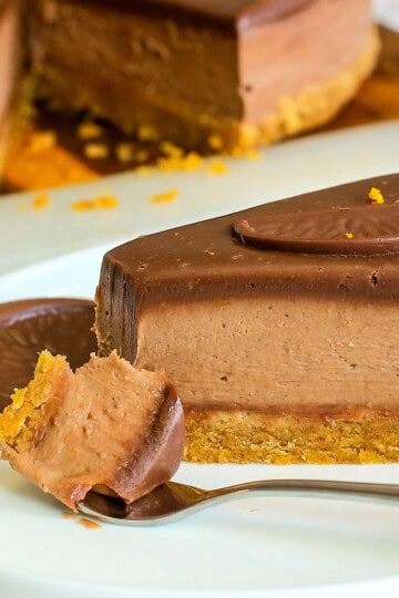 A slice of chocolate orange cheesecake with a biscuit base sits on a white plate, garnished with a chocolate biscuit. A fork holds a bite of the creamy cheesecake in front of the slice.
