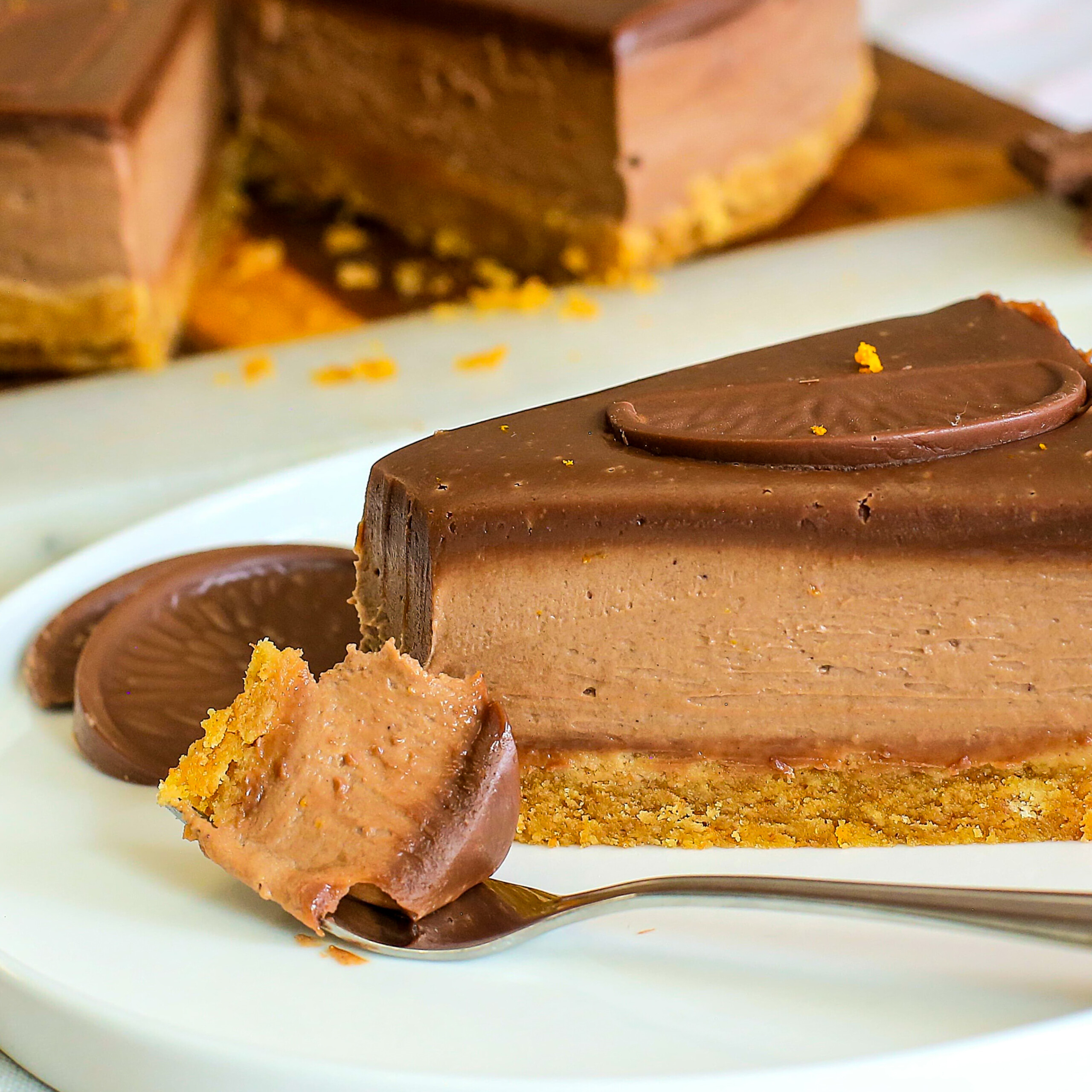 A slice of chocolate orange cheesecake with a biscuit base sits on a white plate, garnished with a chocolate biscuit. A fork holds a bite of the creamy cheesecake in front of the slice.