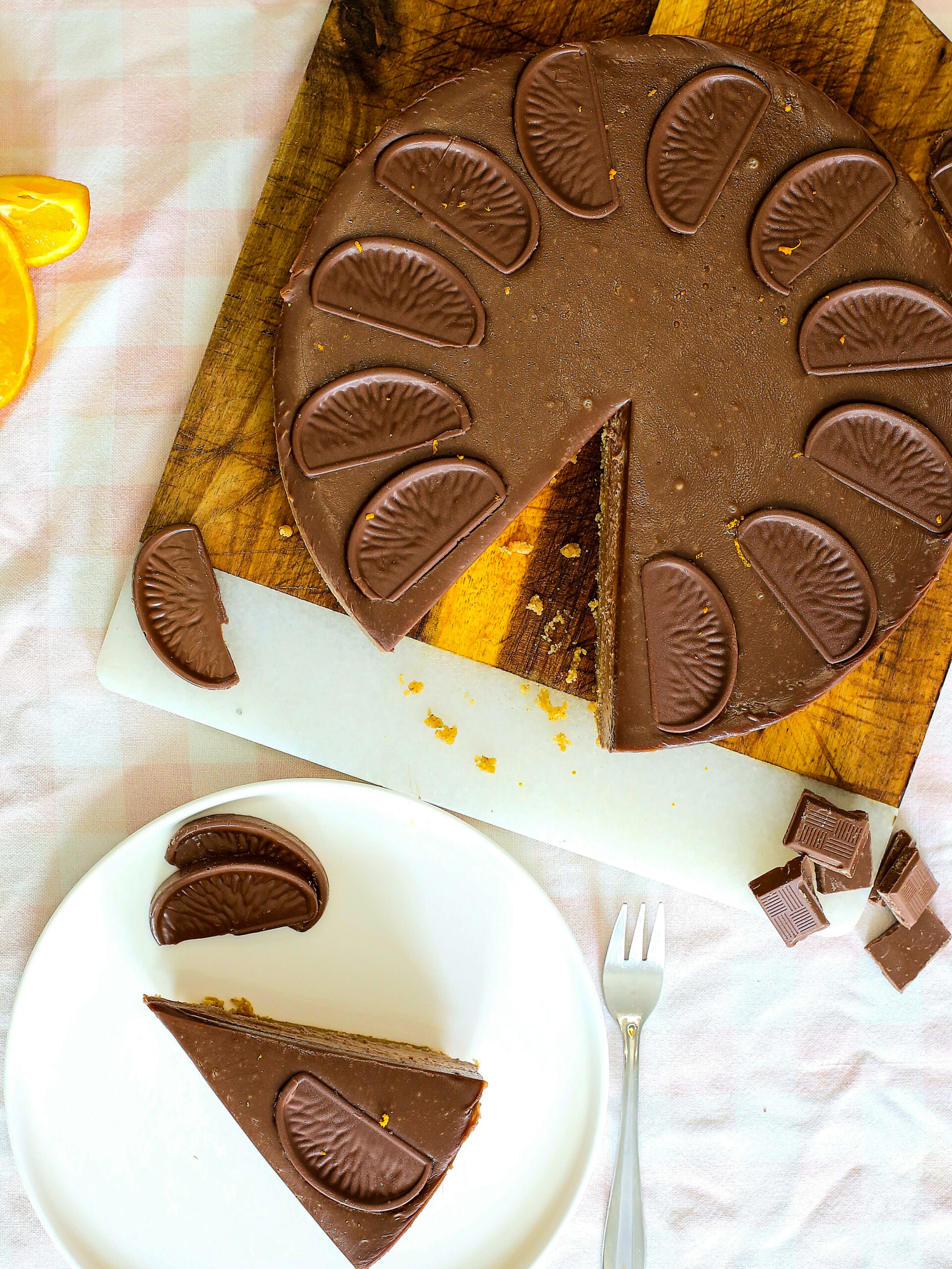 A round chocolate cake topped with chocolate orange segments sits on a chopping board. A slice is cut out and placed on a plate with extra chocolate pieces, alongside a small fork.