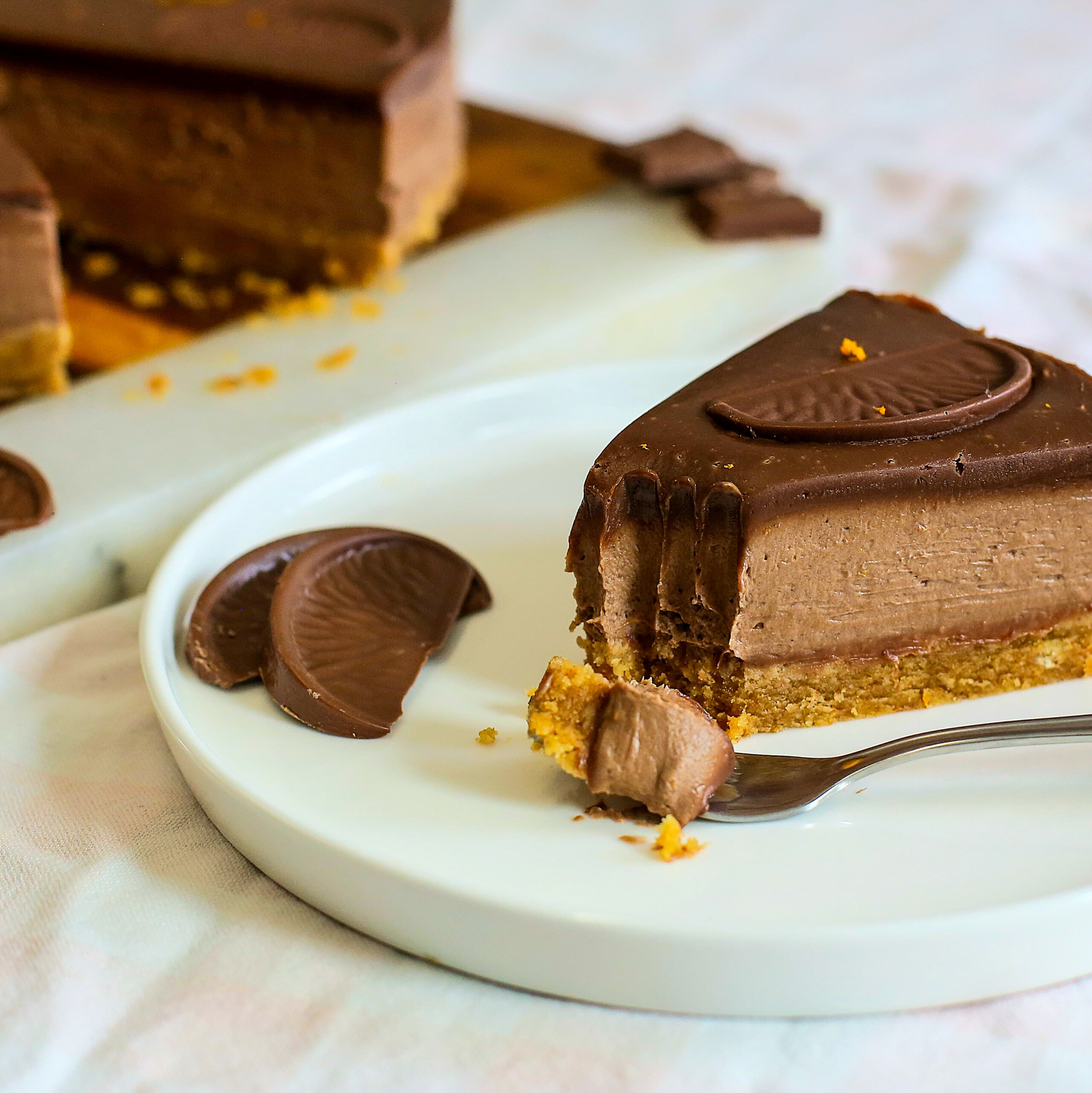 A slice of chocolate orange cheesecake with a biscuit base and chocolate decorations sits on a white plate. A fork holds a bite-sized piece, and more cheesecake is visible in the background.