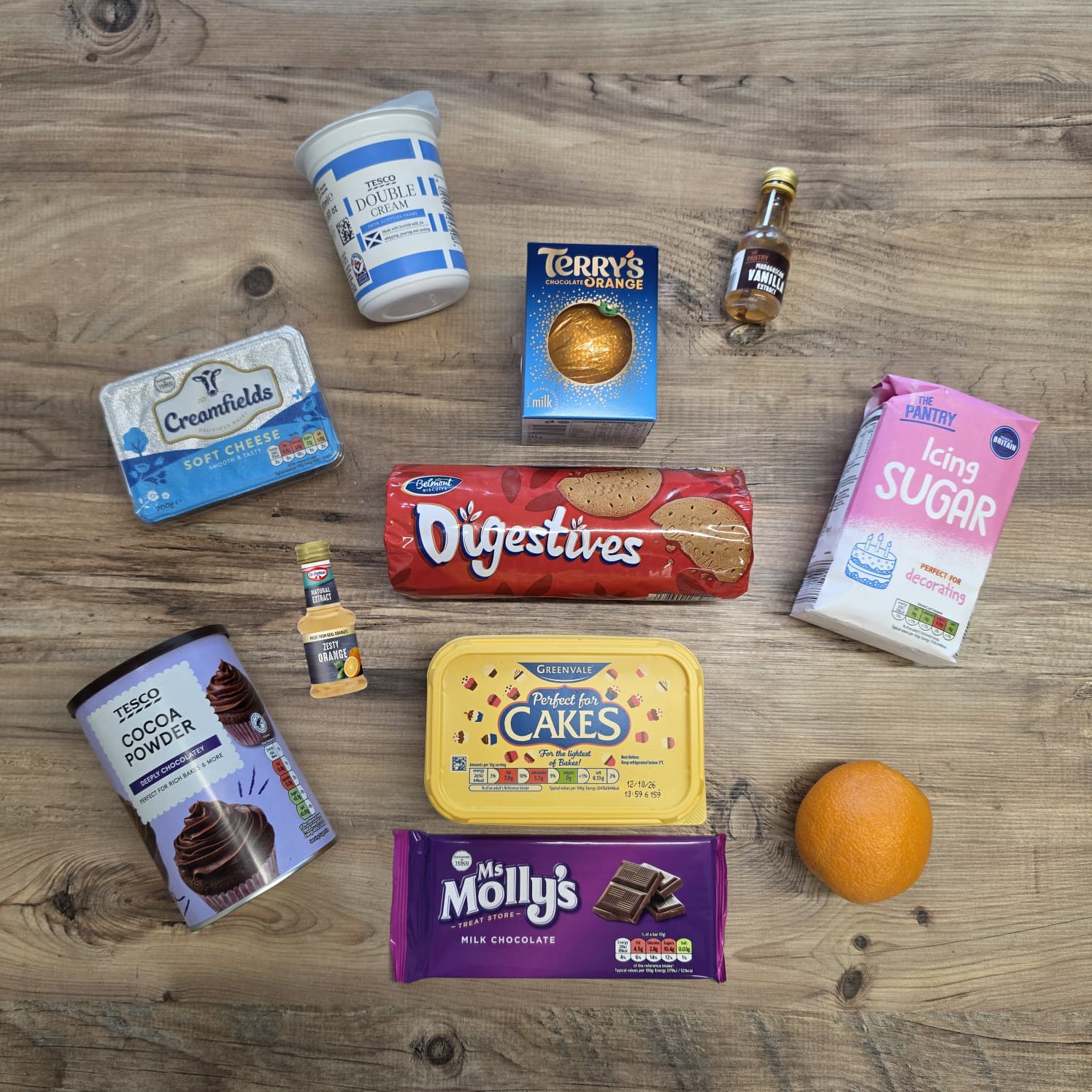 Assorted baking ingredients including cream cheese, chocolate, biscuits, butter, sugar, cocoa powder, orange, and small liqueur bottles are arranged on a wooden surface.