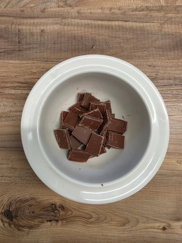 A white bowl filled with small pieces of milk chocolate sits on a wooden surface.