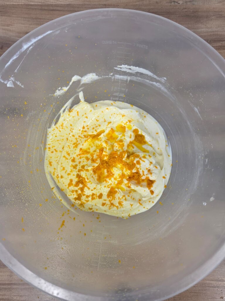 A clear mixing bowl contains a creamy white mixture topped with grated orange zest, sitting on a wooden surface.