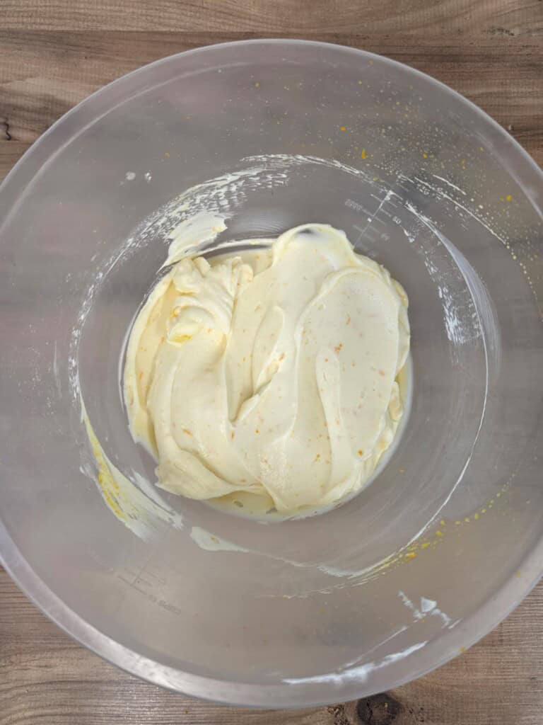 A clear mixing bowl with creamy, pale yellow mixture inside, resting on a wooden surface. The mixture appears smooth with small orange specks throughout.