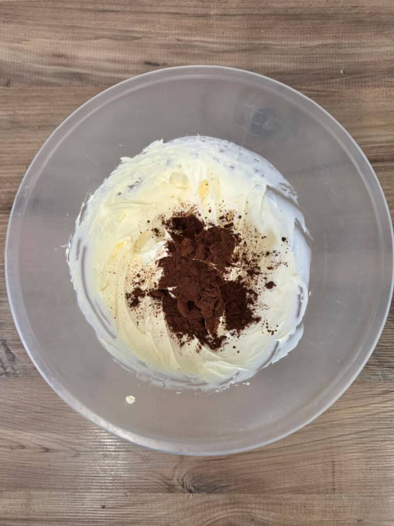 A clear bowl on a wooden surface contains whipped cream cheese or batter with cocoa powder sprinkled on top, ready to be mixed.