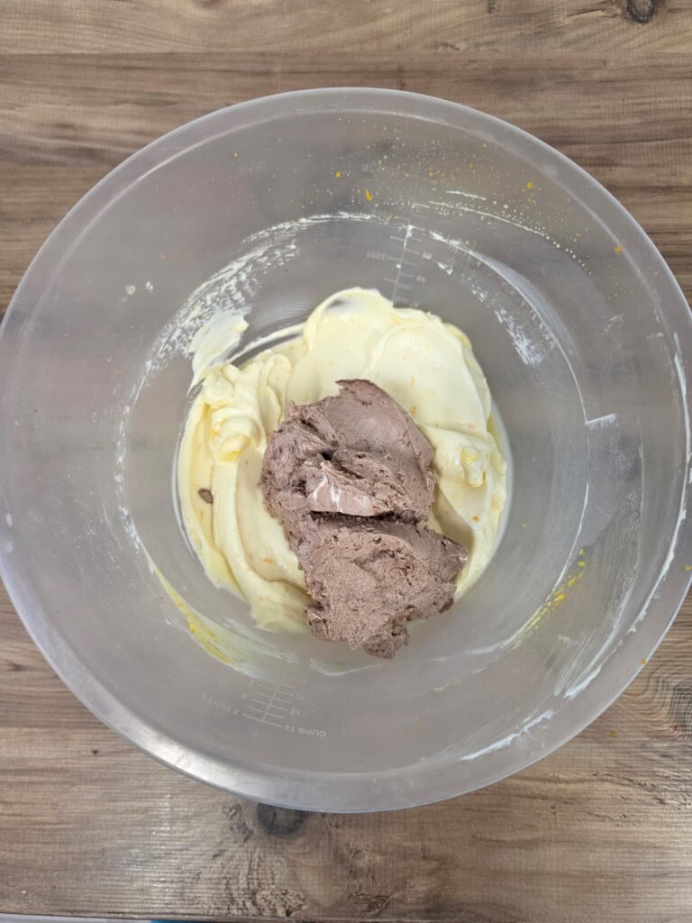 A clear mixing bowl with vanilla and chocolate ice cream sits on a wooden surface, ready to be mixed or served.