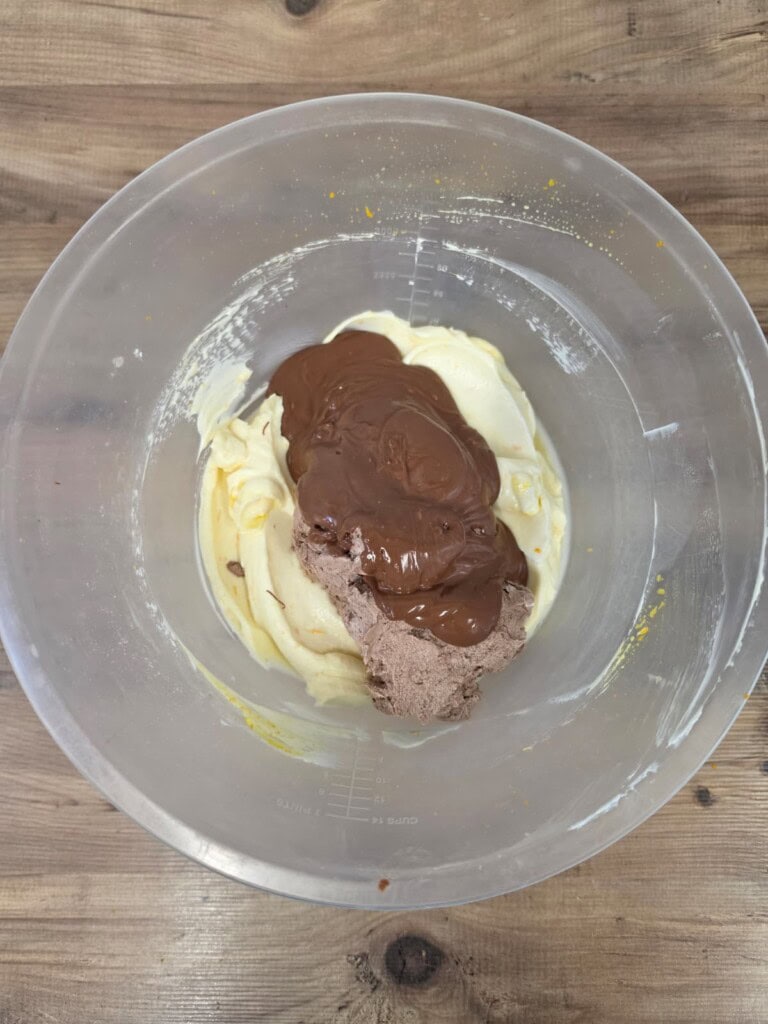 A clear mixing bowl with thick white cream and a dollop of chocolate cream on top, placed on a wooden surface.