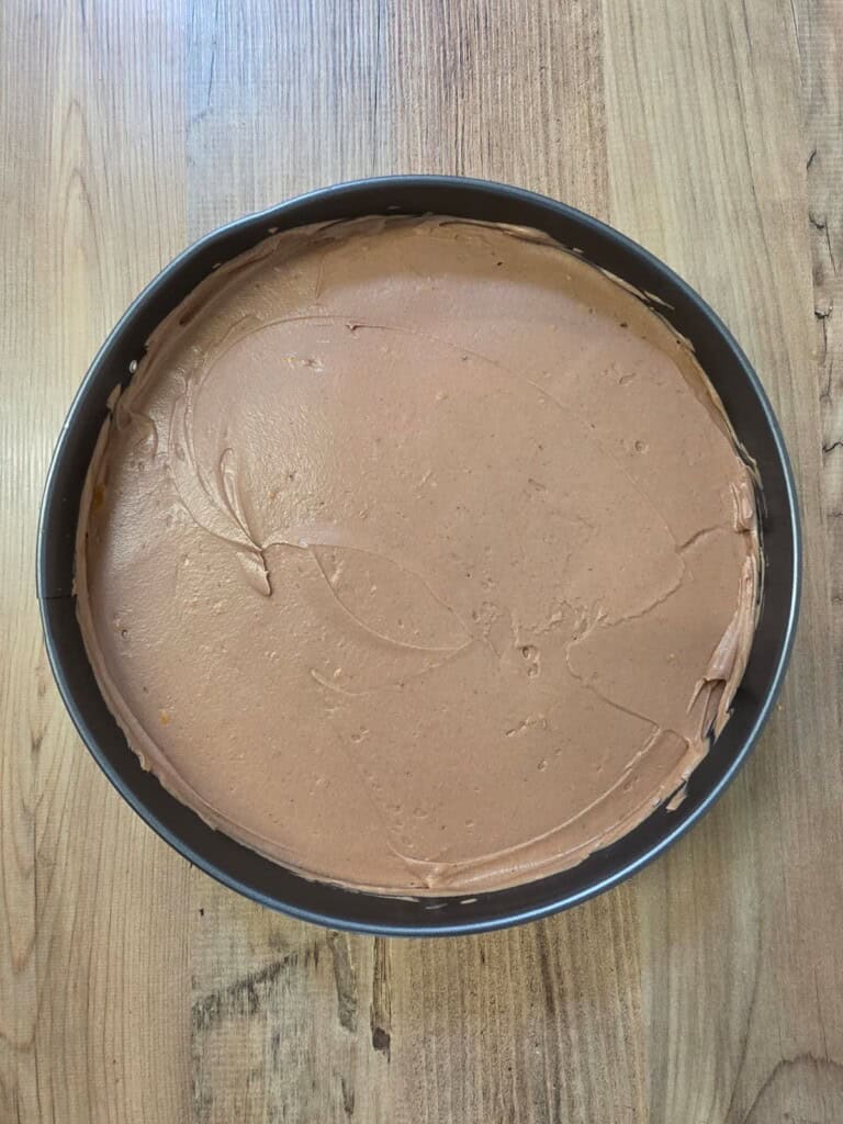 A round tin filled with smooth, light brown batter or mousse, sitting on a wooden surface. The mixture has some swirled marks on its surface.