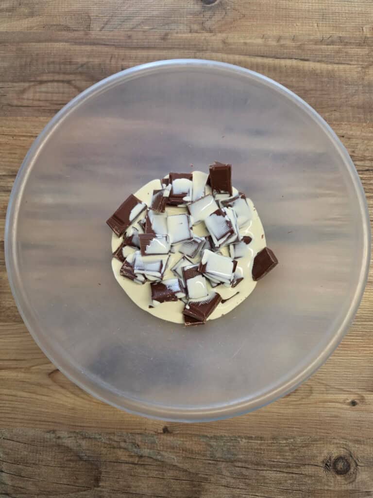 A clear plastic bowl with chunks of milk and white chocolate on a wooden surface.