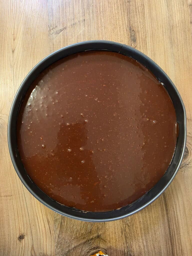 A round baking tin filled with smooth, liquid chocolate cake mixture sits on a wooden surface, ready to be baked.
