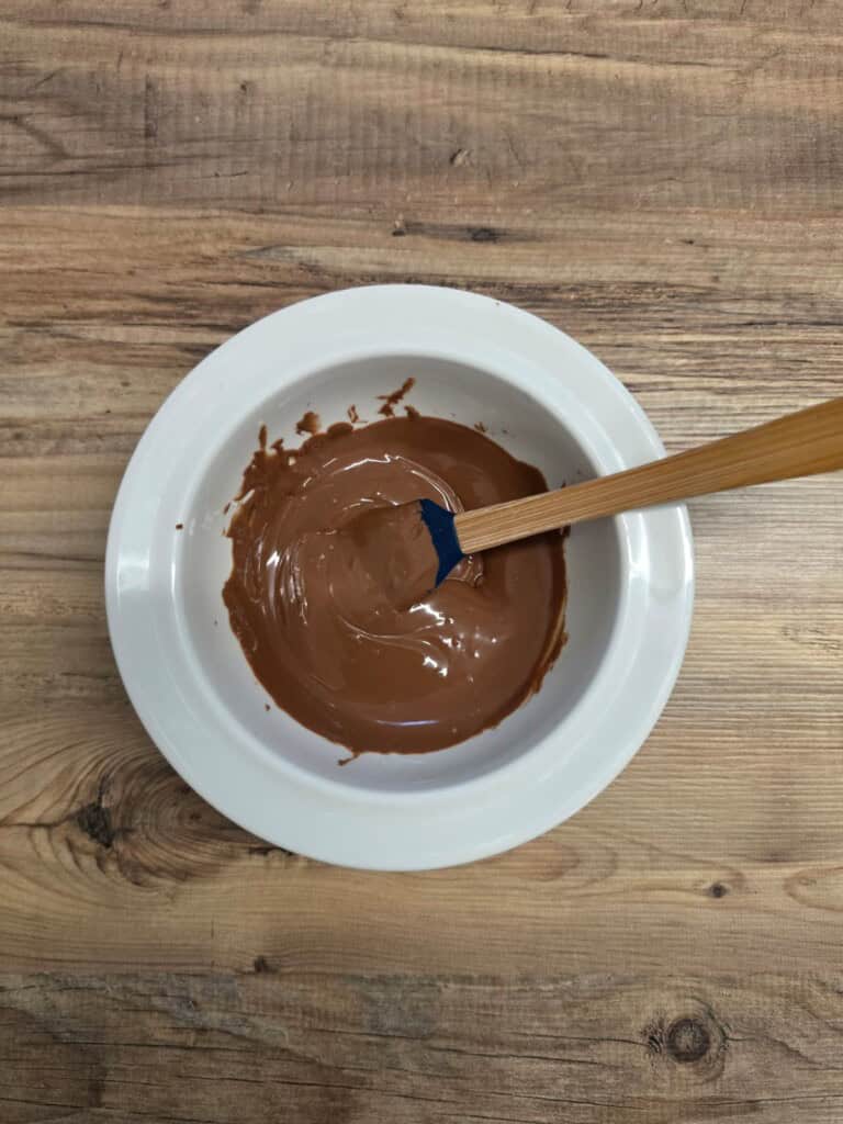 A white bowl filled with melted chocolate is placed on a wooden surface. A blue spatula with a wooden handle is in the bowl, partially covered in chocolate.