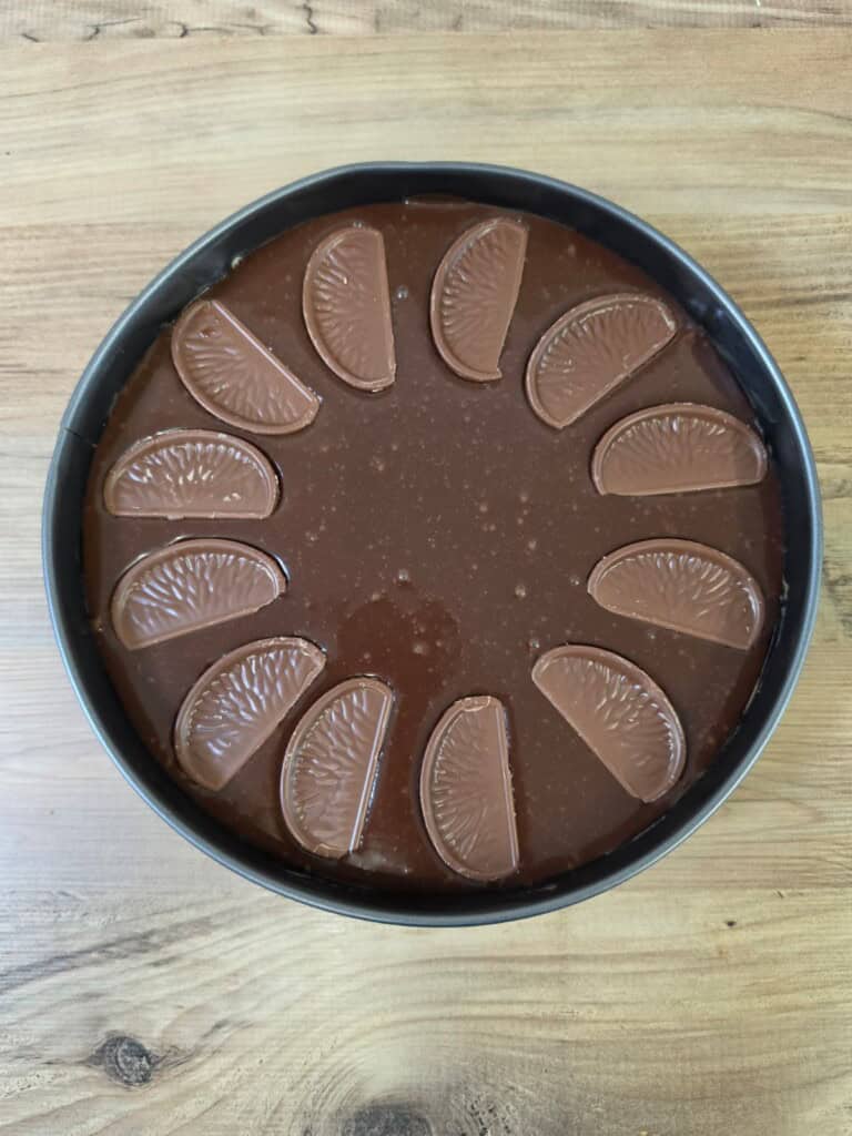 A round chocolate cake topped with a glossy chocolate layer and decorated with twelve chocolate orange segments arranged in a circle, all on a wooden surface.