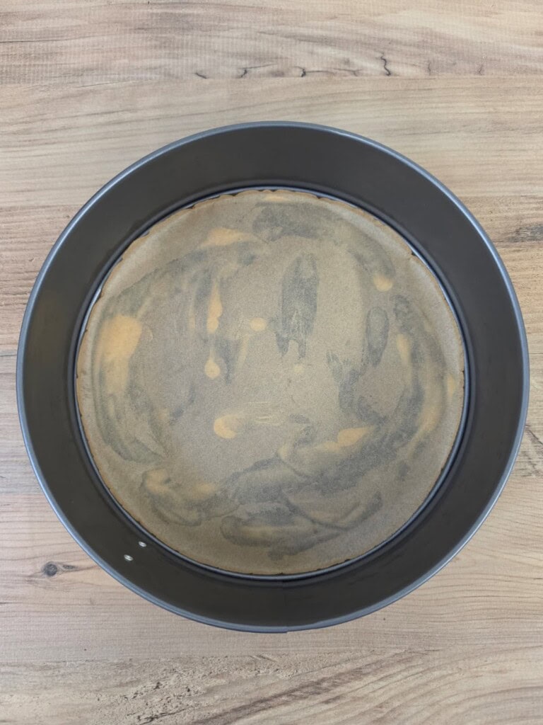 A round springform tin with baking parchment lining the bottom, placed on a wooden surface. The parchment has some light smudges and uneven areas from greasing.