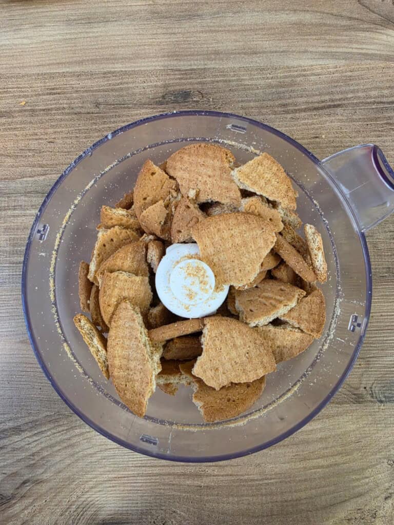 Broken digestive biscuits inside a clear food processor bowl sit on a wooden surface, ready to be blended into crumbs for a recipe.