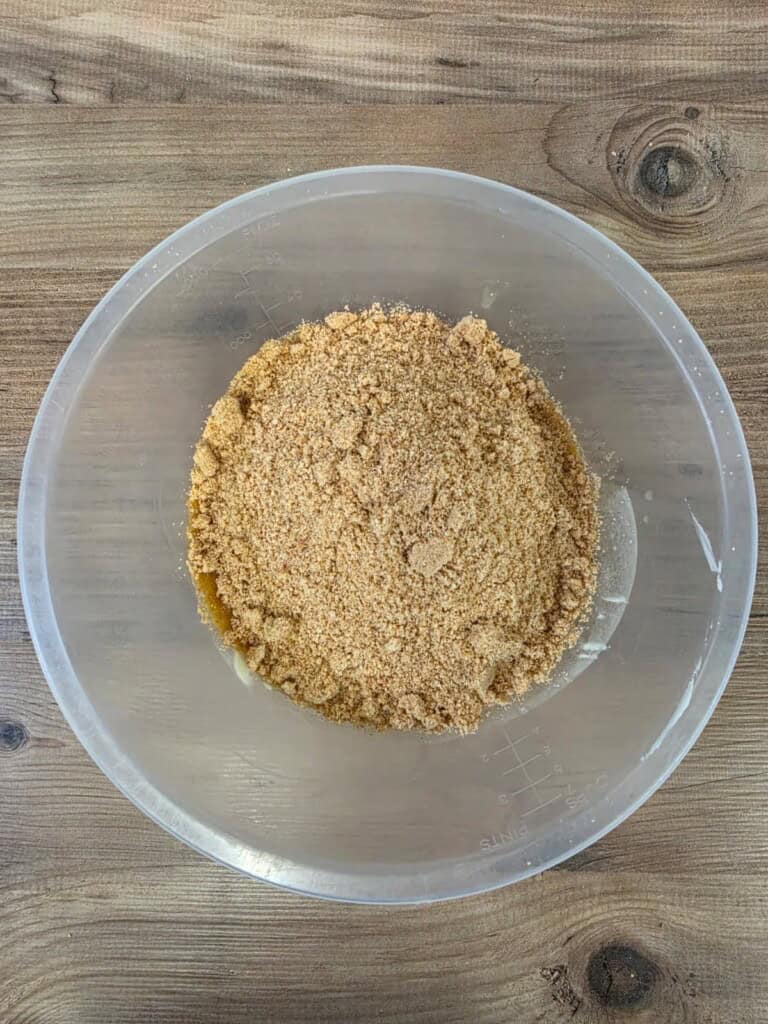A plastic mixing bowl filled with crushed digestive biscuit crumbs sits on a wooden surface, ready to be used as an ingredient for baking.