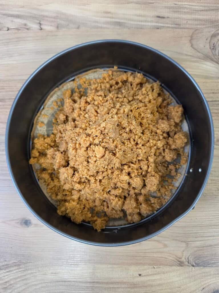 A round metal cake tin on a wooden surface, filled with crumbled biscuit or digestive biscuit base, not yet pressed down or baked.