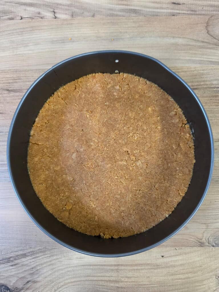 A round baking tin filled with a smooth, pressed biscuit or digestive biscuit base, sitting on a wooden surface, ready for baking or filling.