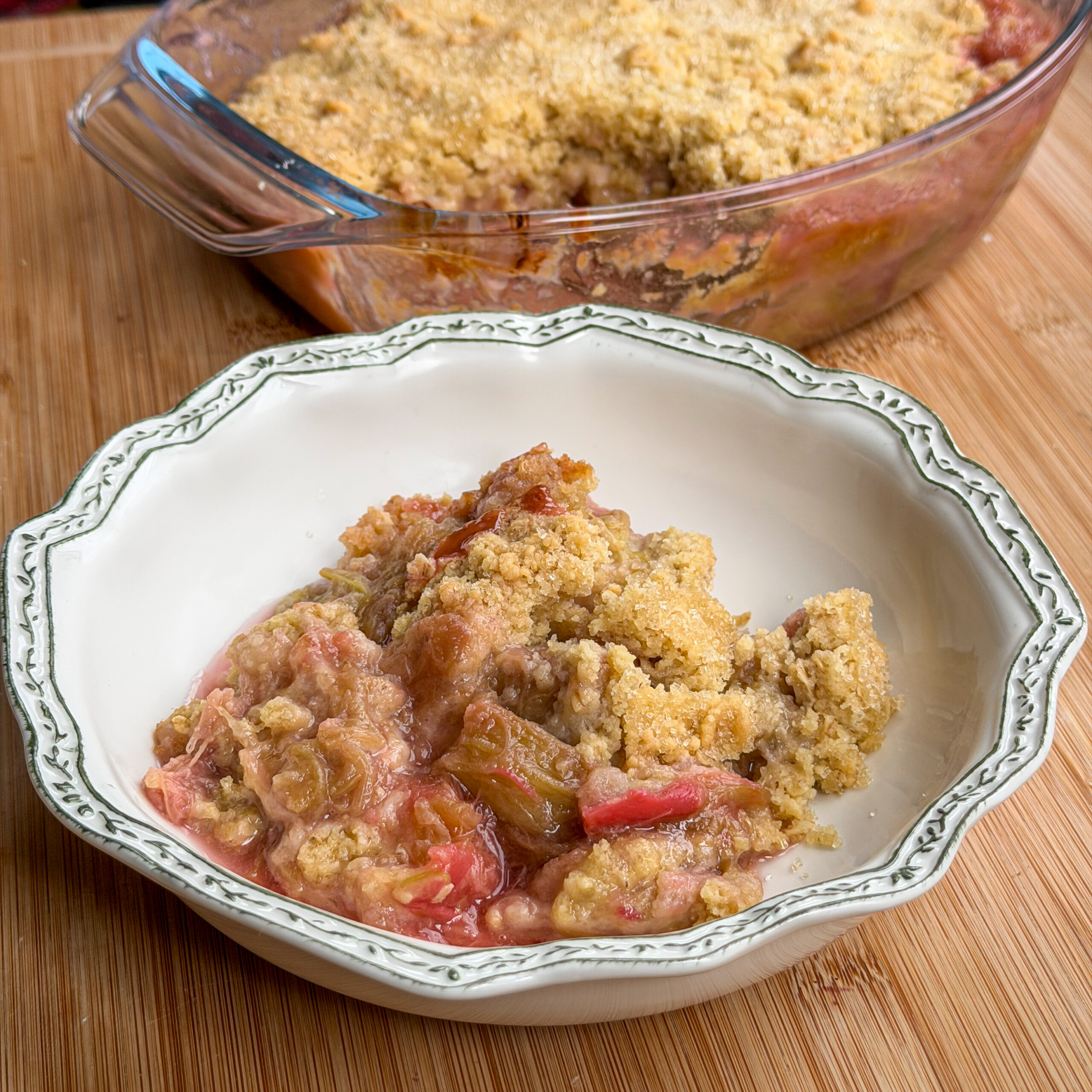 A serving of rhubarb crumble with a golden crumbly topping sits in a decorative white bowl. The glass baking dish with more crumble is in the background on a wooden surface.