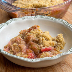 A serving of rhubarb crumble with a golden, crumbly topping sits in a decorative white bowl. A glass baking dish with more of the pudding is in the background on a wooden surface.