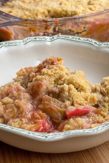 A serving of rhubarb crumble with a golden, crumbly topping sits in a decorative white bowl. A glass baking dish with more of the pudding is in the background on a wooden surface.