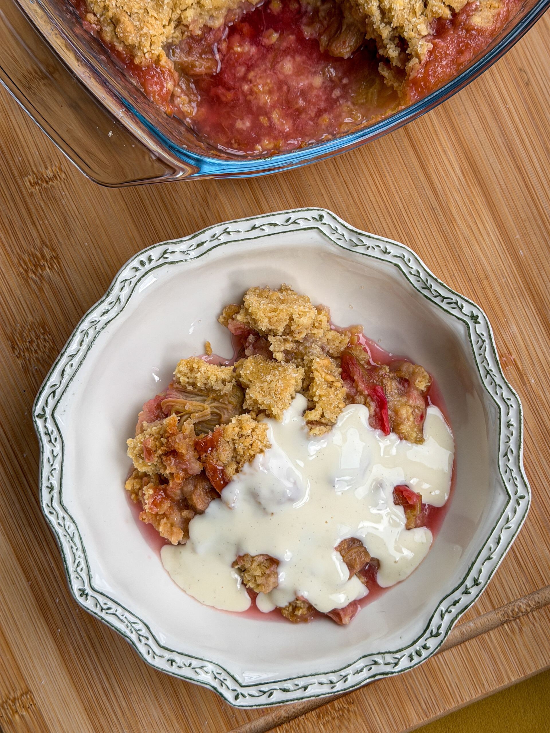 A bowl of fruit crumble pudding with cream poured on top sits on a wooden surface next to a baking dish containing more of the crumble.