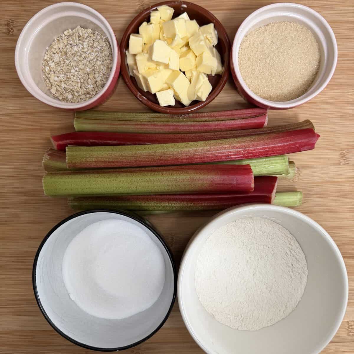 Rhubarb stalks with bowls of oats, diced butter, brown sugar, white sugar, and flour on a wooden surface, arranged neatly for baking preparation.