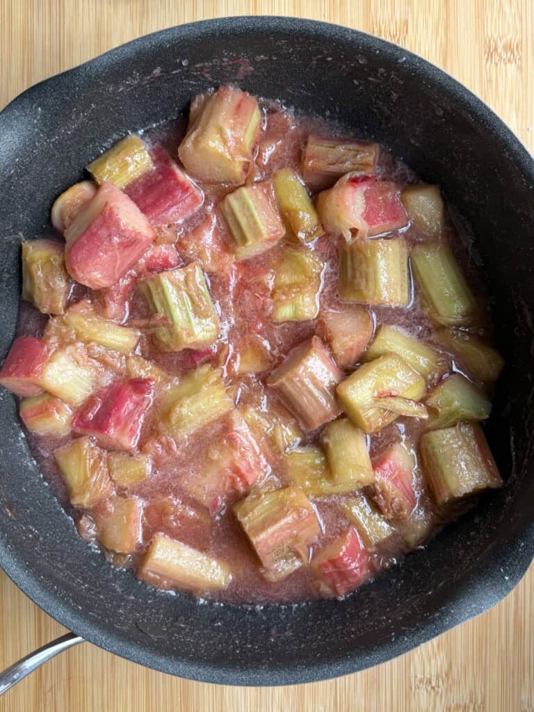 Chunks of cooked rhubarb in a black saucepan, sitting on a light wooden surface. The rhubarb pieces are soft and surrounded by a pinkish juice.