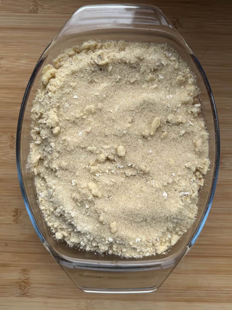 A glass baking dish filled with an even layer of crumbly, light brown topping, likely an unbaked crumble or crisp pudding, sits on a wooden surface.