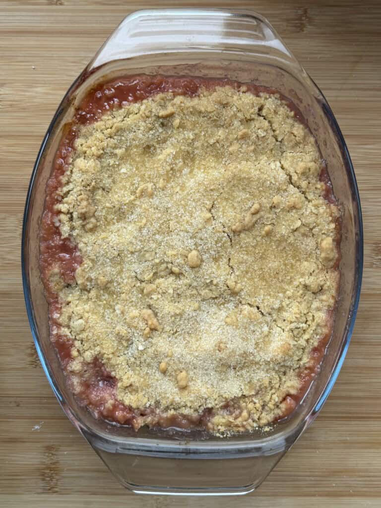 A glass baking dish filled with a golden-brown fruit crumble sits on a wooden surface. The crumble topping appears crisp and slightly uneven, with some red fruit filling visible at the edges.