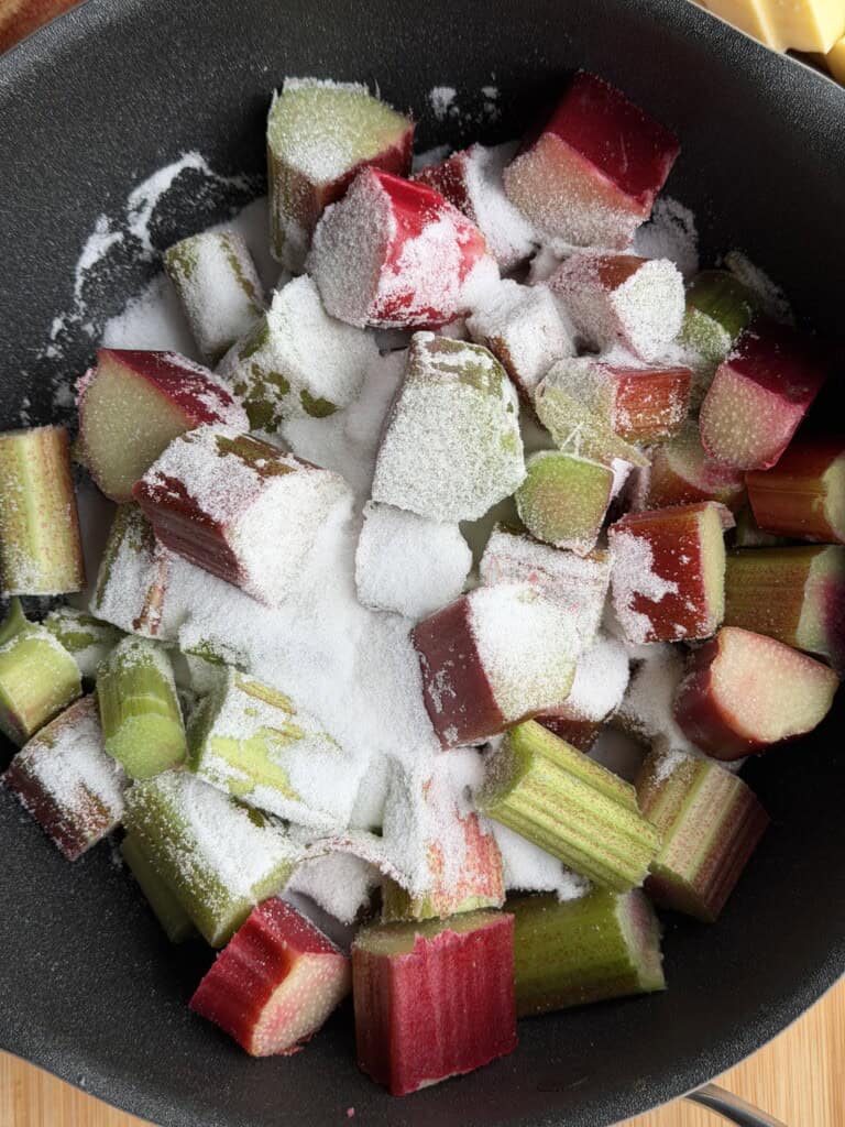 Chopped rhubarb pieces in a saucepan are sprinkled with white caster sugar, ready for cooking or baking.